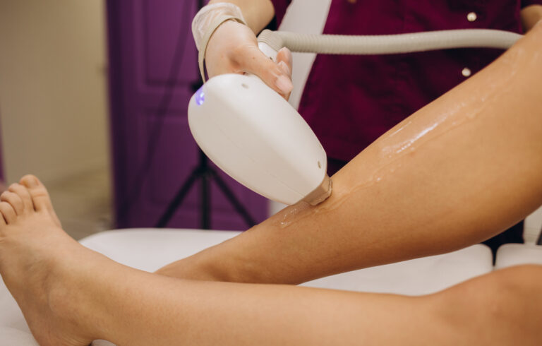 A woman is lying down while a technician holds a device against her leg for laser hair removal in Frisco.